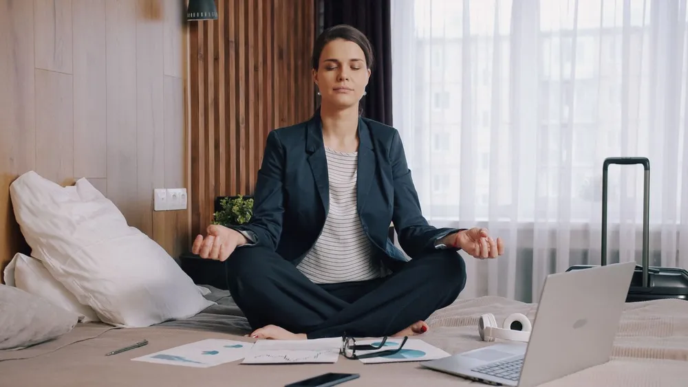 Mulher de blazer meditando sentada, ao lado do notebook e dos papéis de trabalho